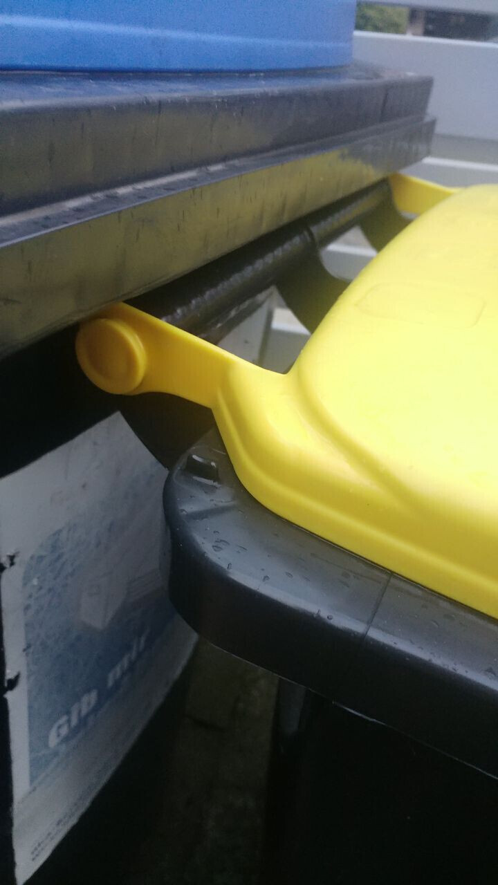 Nahaufnahme eines gelben Deckelverschlusses an einem Müllcontainer; schwarzer Deckel unten.