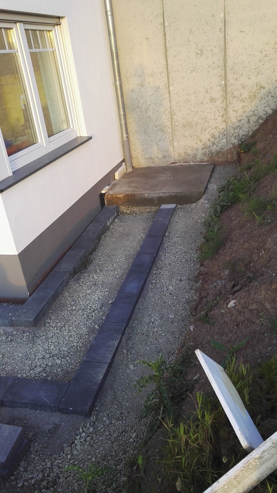 Außenansicht eines Hauses mit Fenster; dunkle Pflastersteine bilden Weg neben Kies und Türstufe.