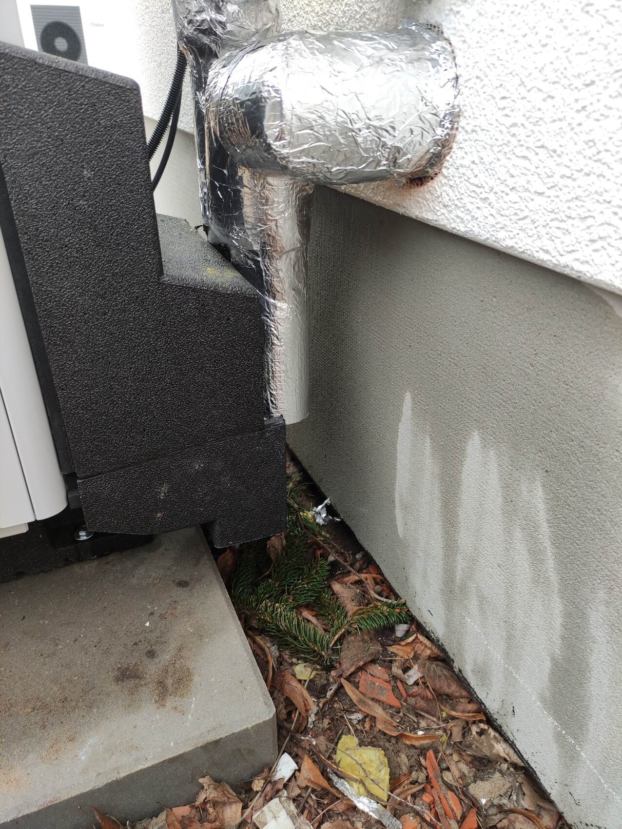 Außengerät einer Klimaanlage mit isoliertem Aluminium-Rohr vor grauer Wand; Laub am Boden.