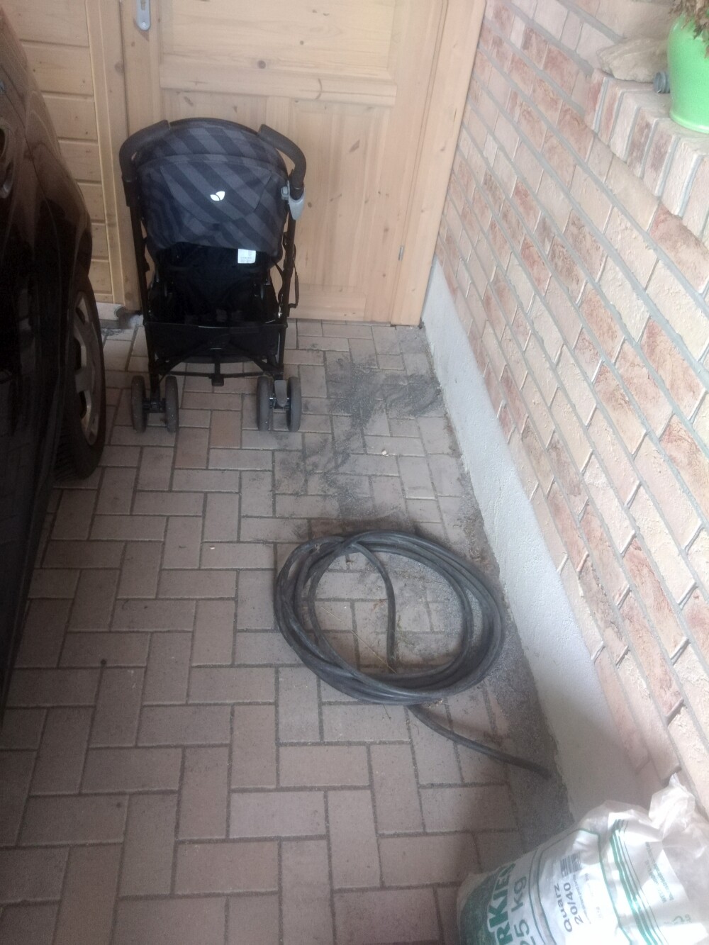 Kinderwagen vor Holztür, gepflasterter Boden, neben aufgerolltem Schlauch an der Wand.