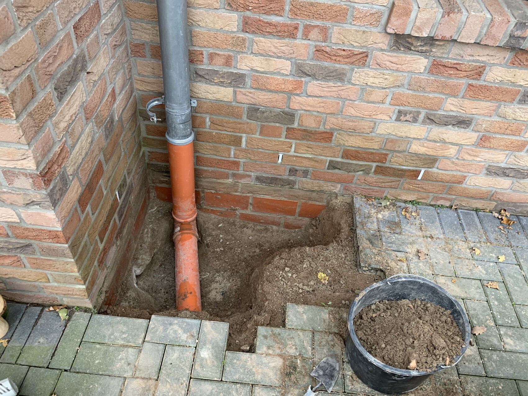 Orangefarbene und graue Abflussrohre führen in Erdgrabung neben der Hauswand; Eimer mit Erdreich.