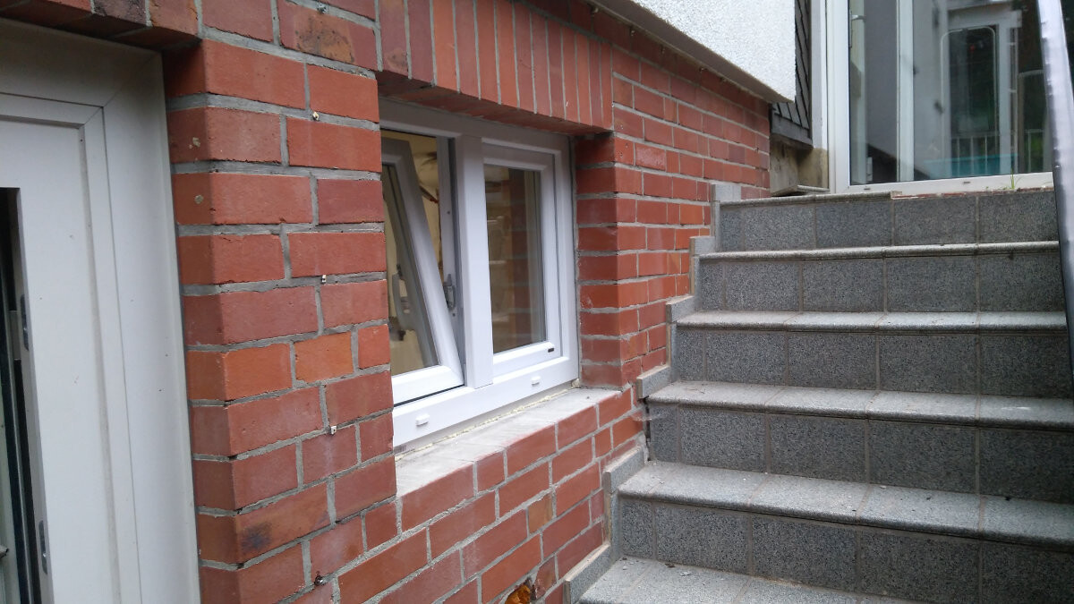 Außenansicht eines Hauses: rote Backsteinwand, weißes Fenster; rechts graue Fliesenstufen.