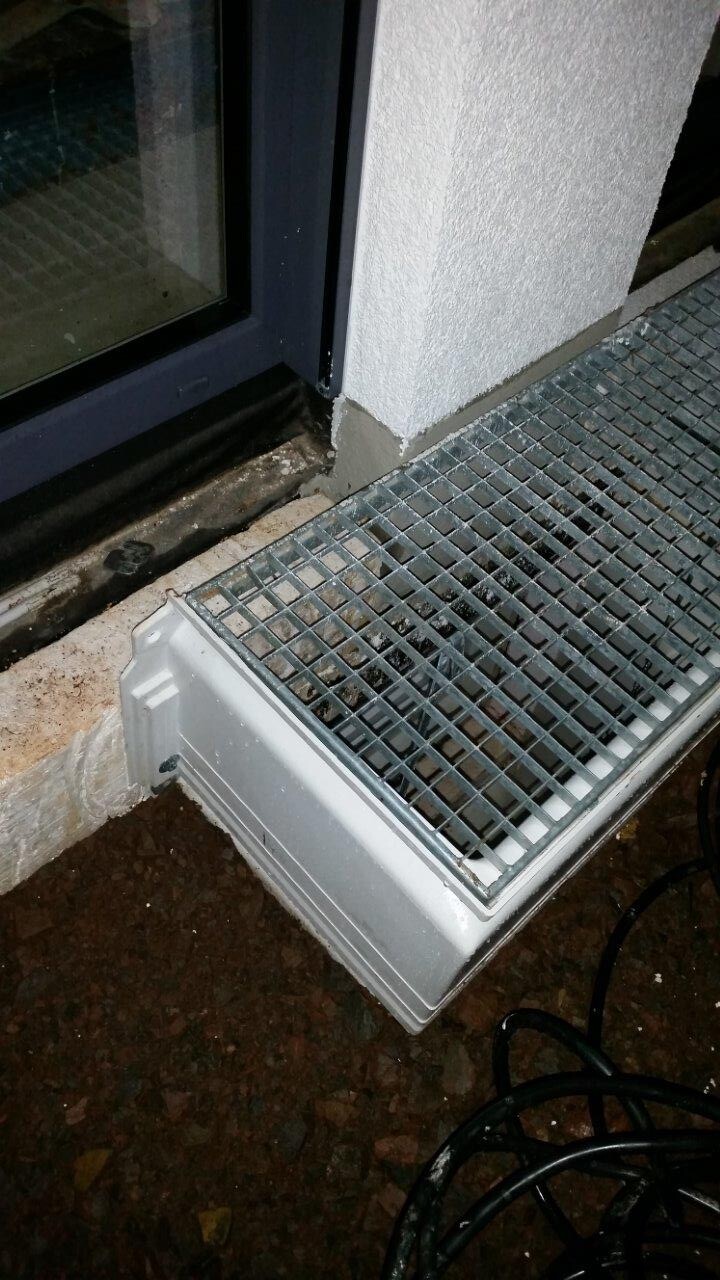 Schräg liegende Gitterrostabdeckung vor einer Glasöffnung neben weißer Wand; Kabel am Boden.