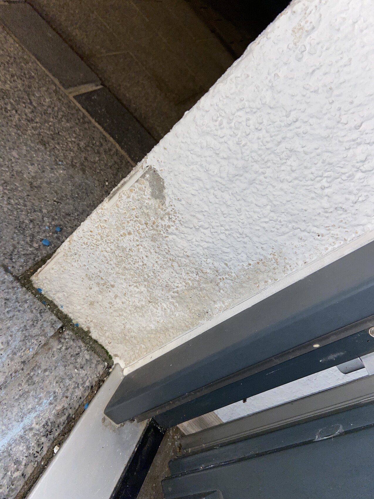 Türschwelle und grauer Metallrahmen vor einer weißen, rauen Wand; Steinboden davor.