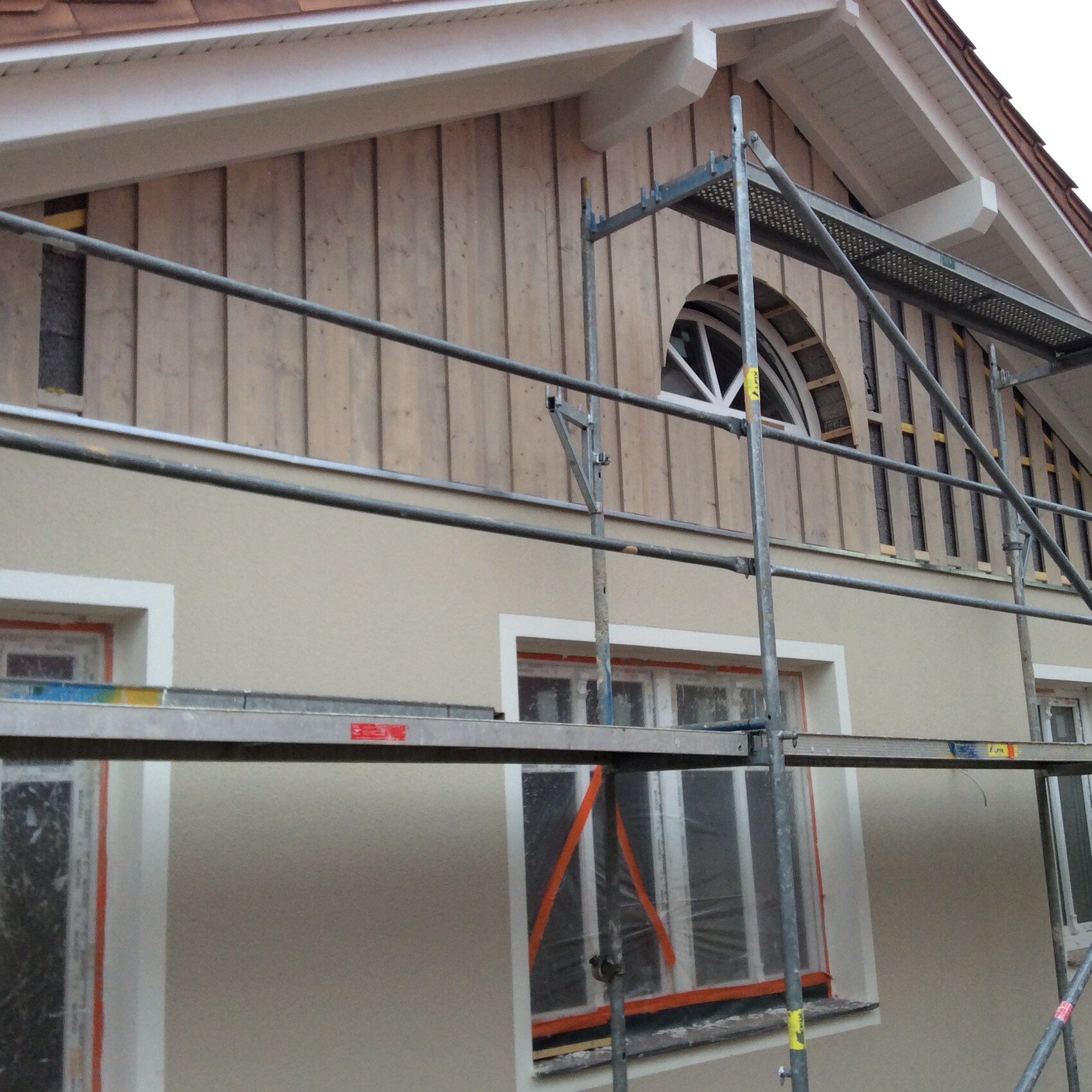 Außenansicht eines Hauses unter Renovierung mit Gerüst, Holzverkleidung und Rundbogenfenster.