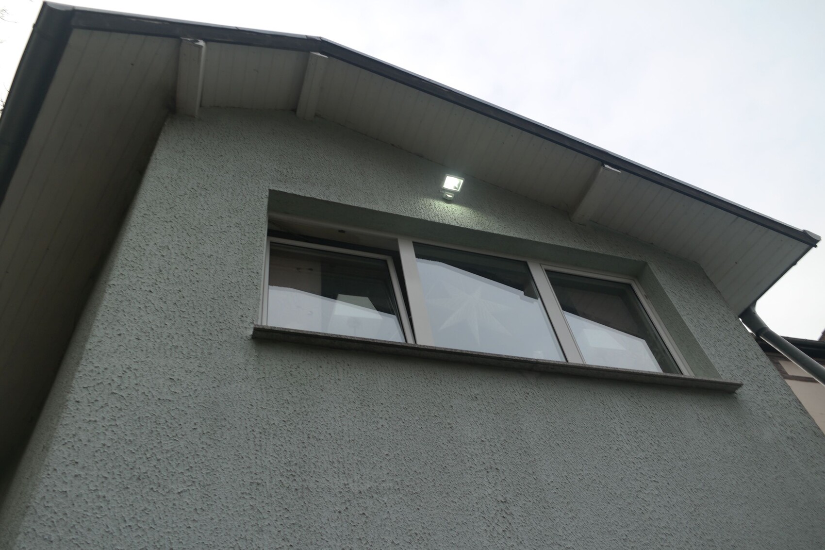 Graue Putzfassade mit großem Fenster und Außenleuchte unter dem Dachüberstand.