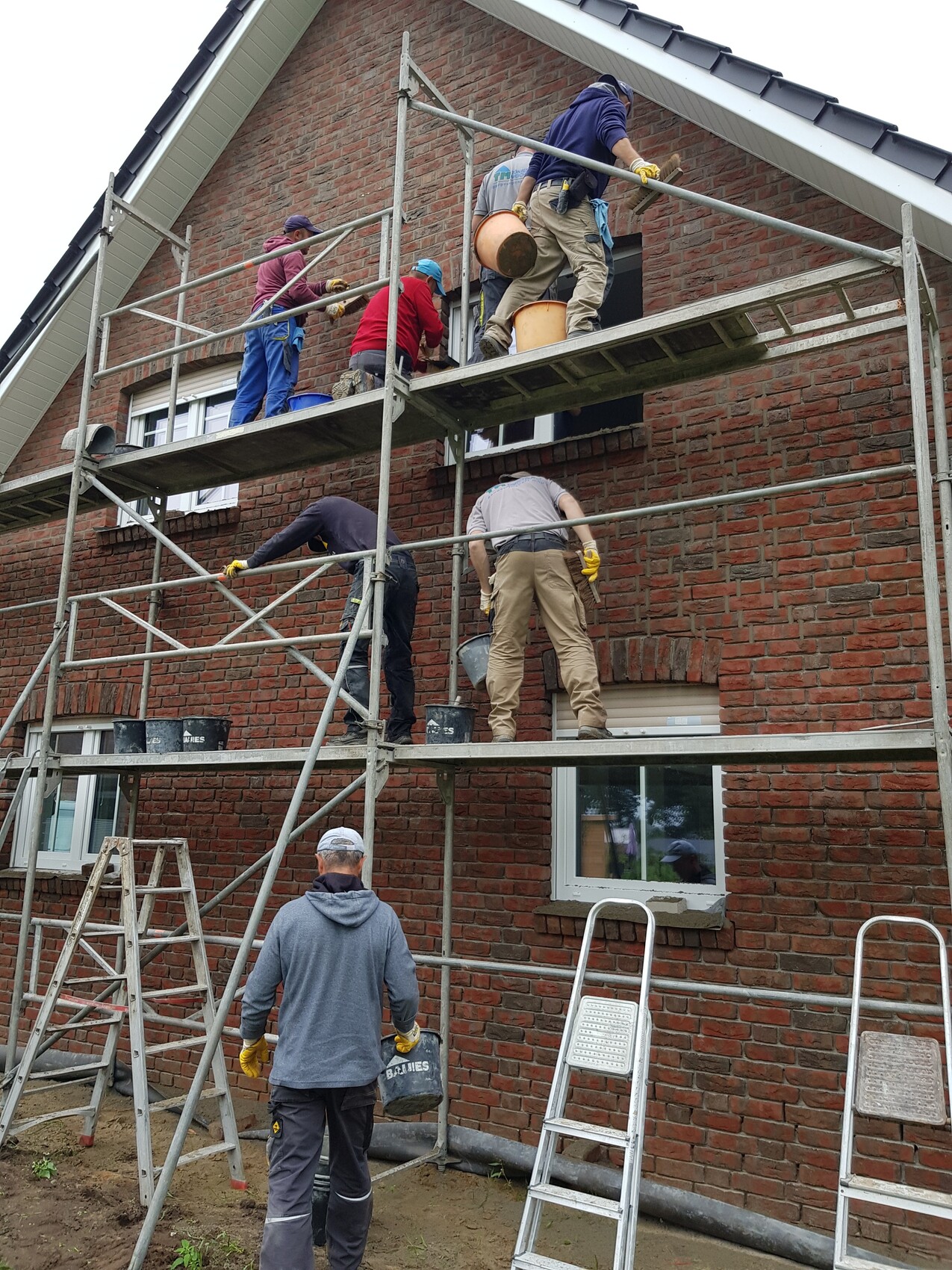 Mehrere Bauarbeiter auf Gerüst arbeiten an roter Backsteinfassade eines Hauses; Eimer bereit.