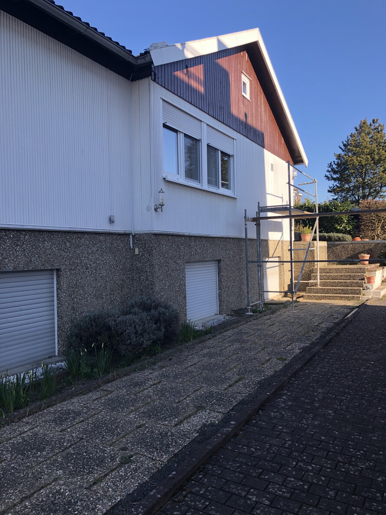 Zweifamilienhaus mit weißer Fassadenverkleidung, braunem Giebel, Baugerüst und gepflastertem Vorplatz.