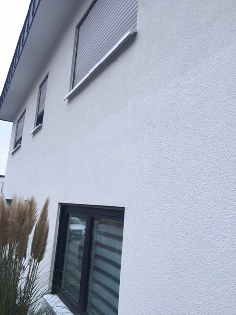 Weiße Außenwand eines Gebäudes mit Rollladenfenstern und bodentiefem Fenster im Erdgeschoss