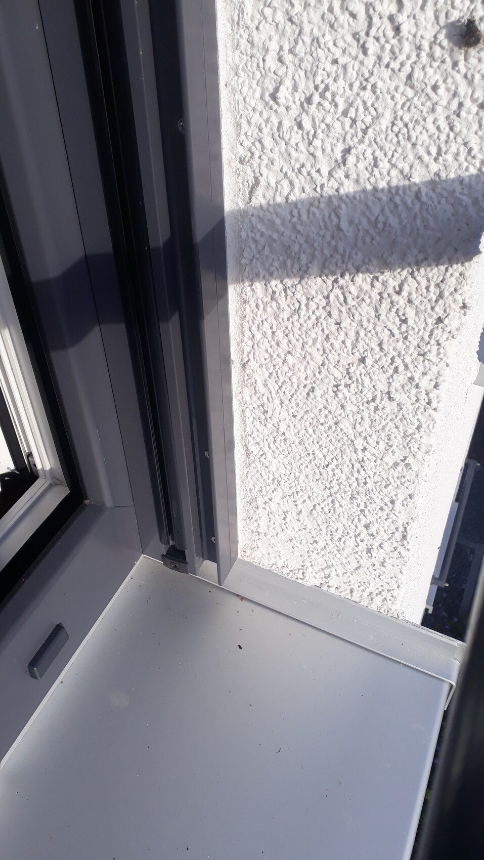 Außenfensterrahmen aus Metall neben rauer weißer Putzwand, Fensterbank im Vordergrund.