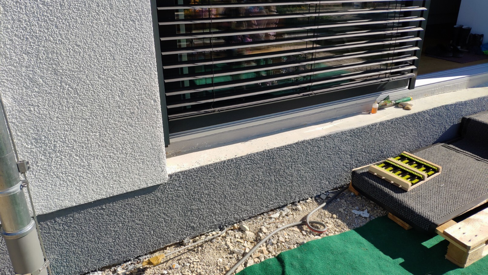 Außenansicht: Fenster mit Jalousien an grauer Wand; Werkzeuge und Bürsten liegen auf einer Matte.