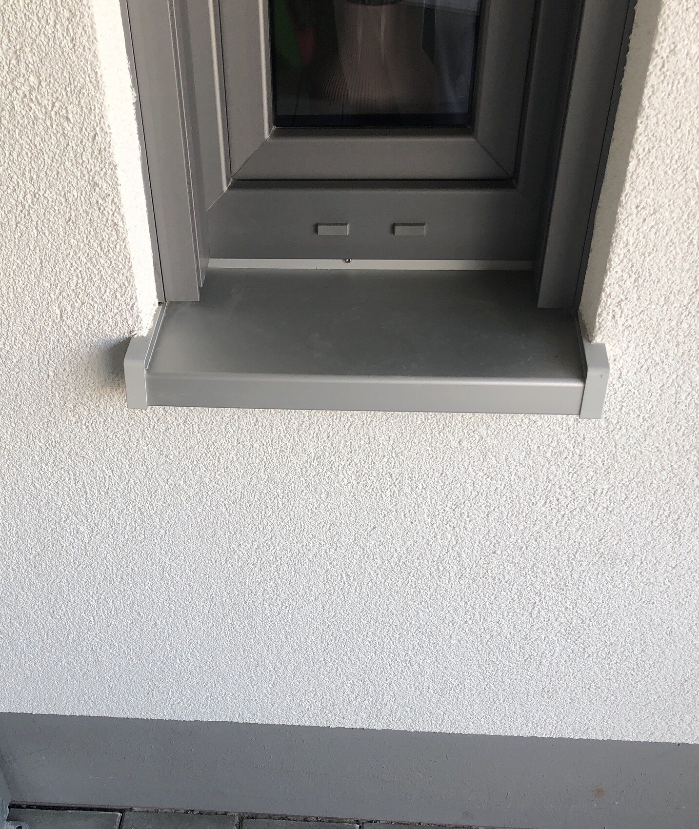 Außenansicht eines grauen Metallfensterrahmens mit Fensterbank vor einer grob texturierten weißen Wand