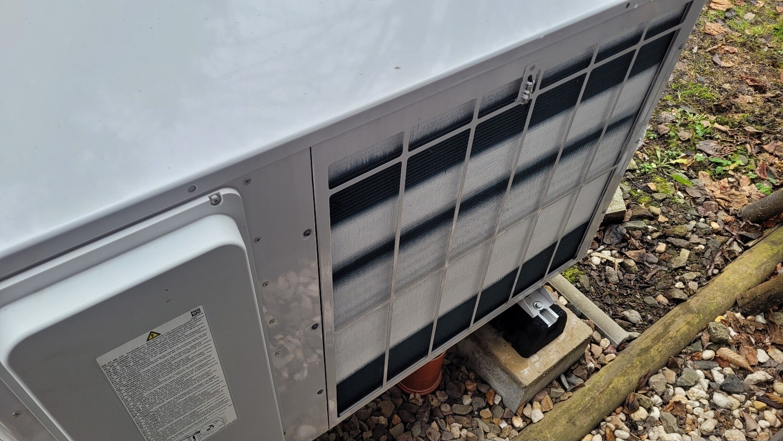 Außenkondensator einer Klimaanlage mit Schutzgitter, auf Kiesboden montiert.