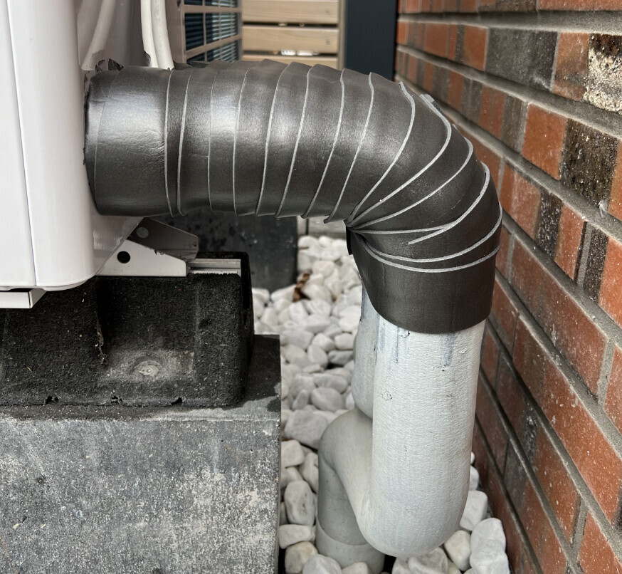 Außengerät einer Klimaanlage mit flexiblem, isoliertem Schlauch und Metallrohr an Ziegelwand.