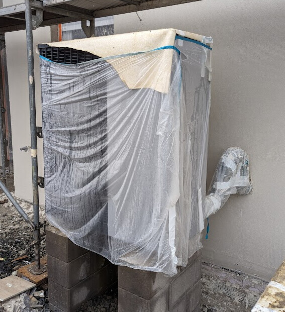Außen-Klimagerät mit Plastikfolie eingewickelt, steht auf Betonblöcken vor der Hauswand.