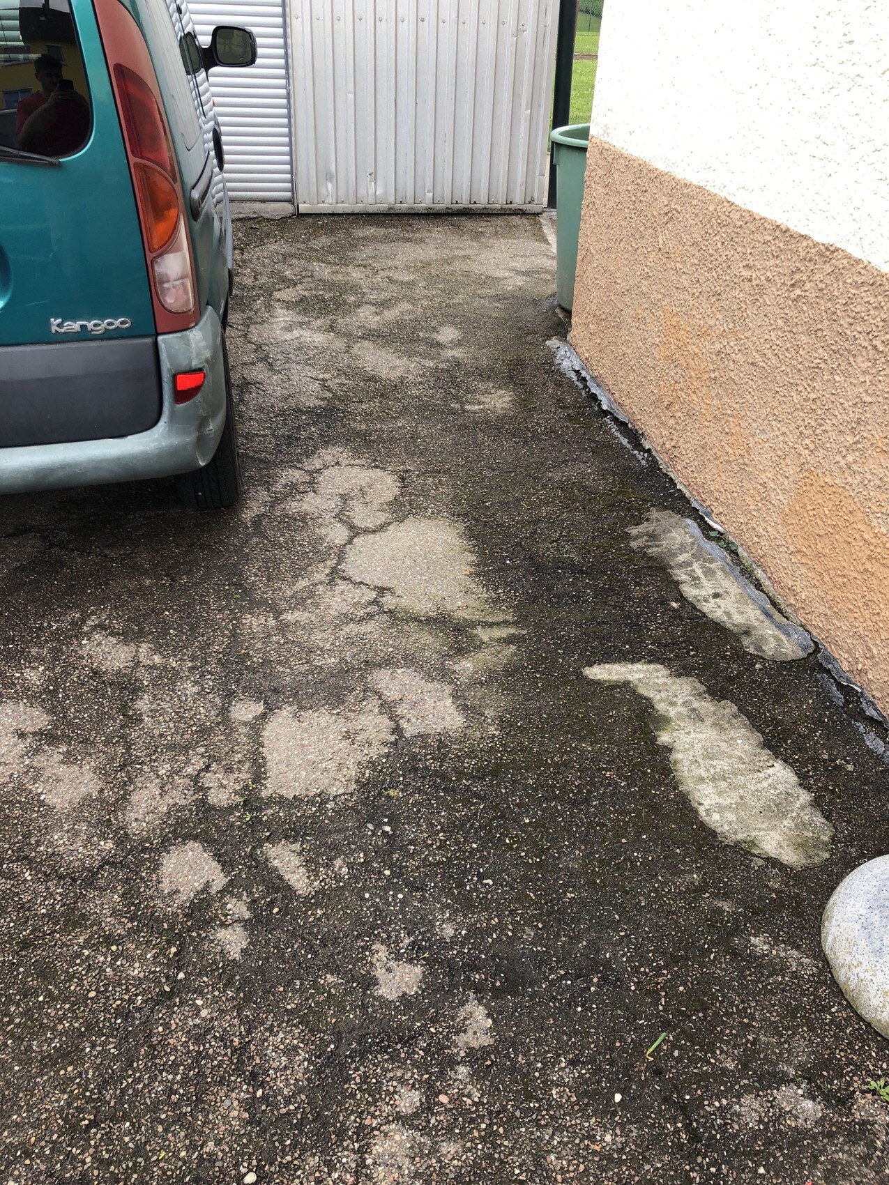 Blauer Kangoo-Van parkt links, unebener Asphalt mit Pfützen neben einer beigen Außenwand.