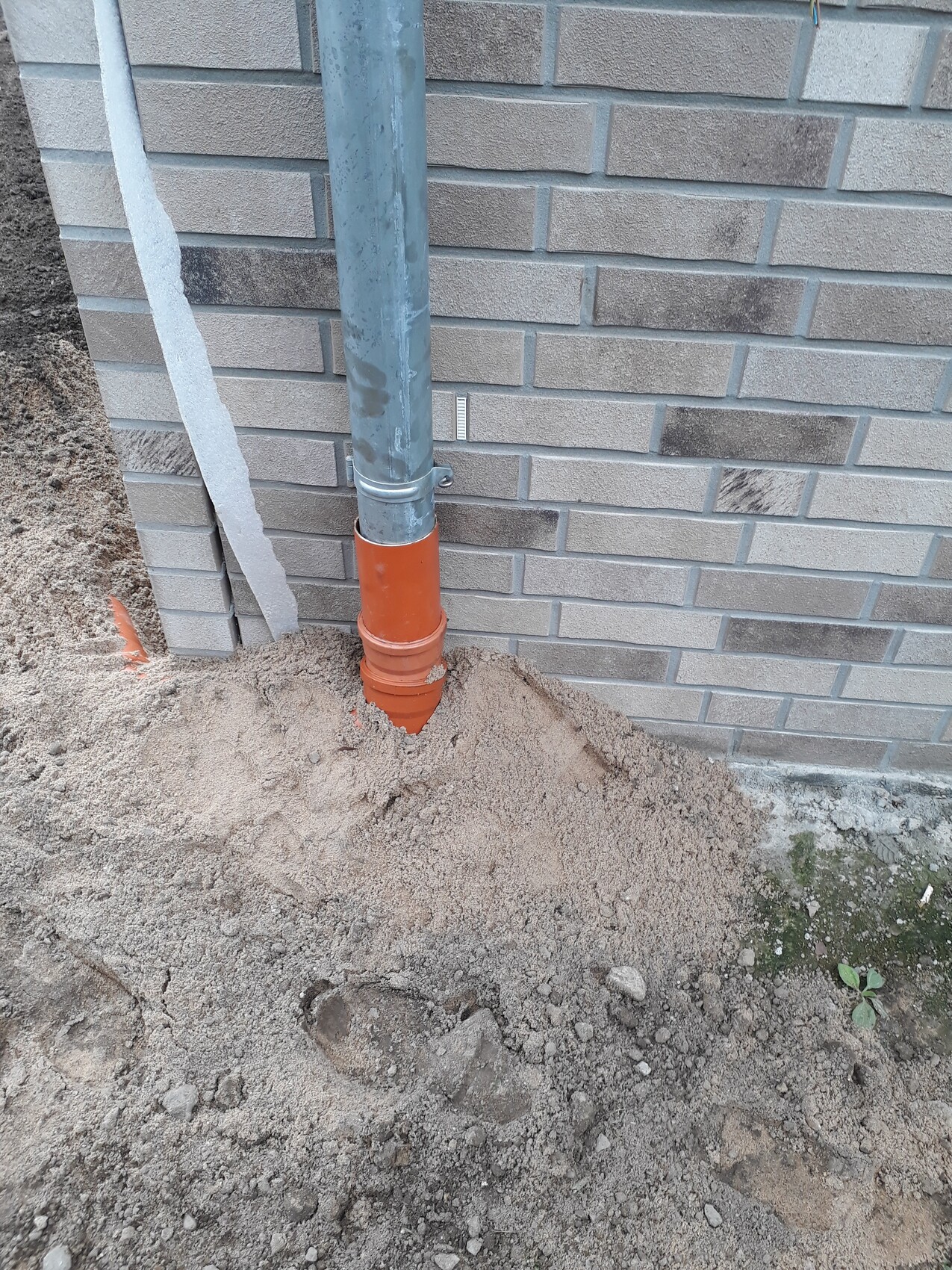 Fallrohr aus Metall mündet in orange PVC-Rohr im Erdreich neben einer Ziegelwand, Sand bedeckt.