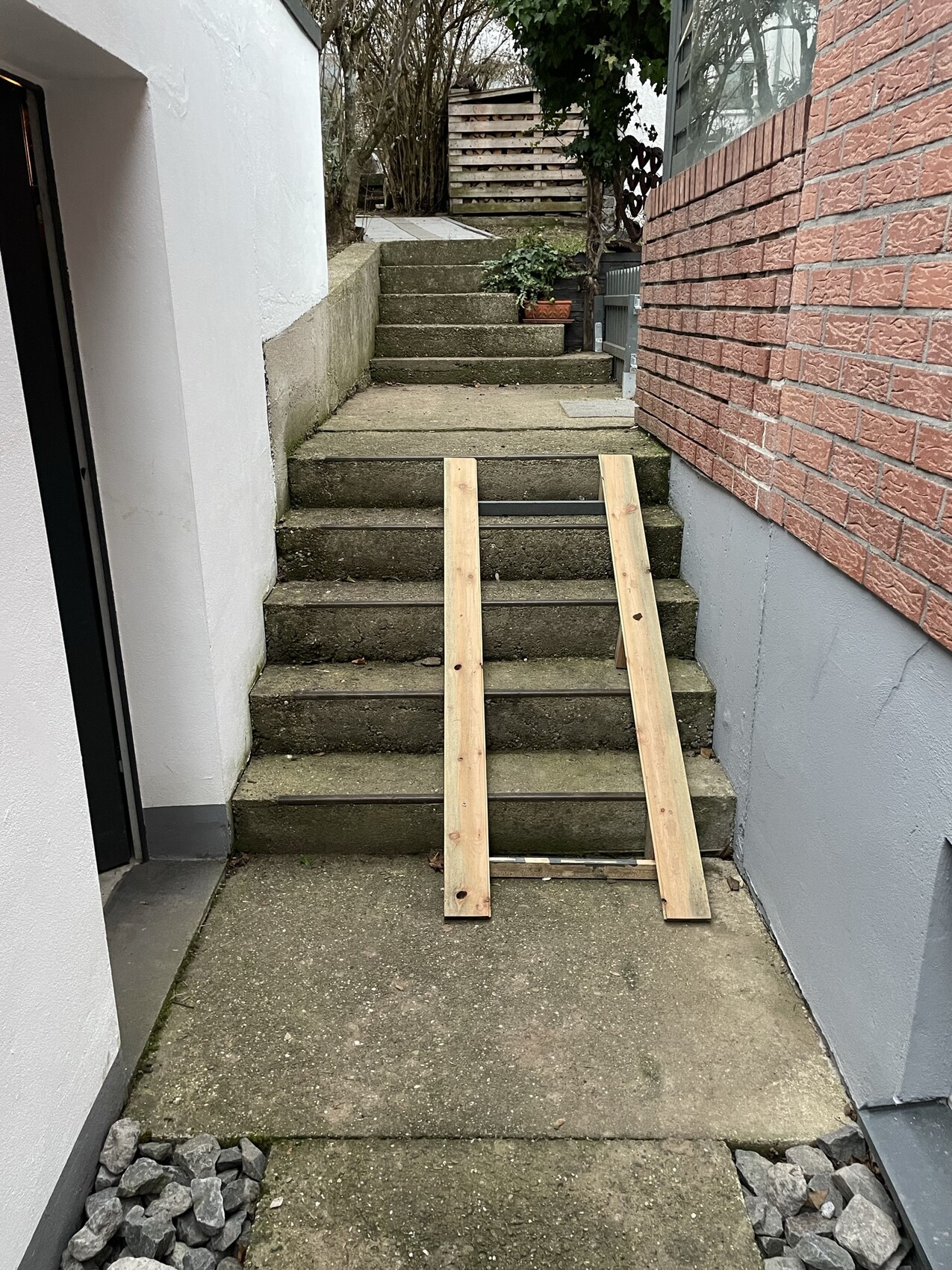 Provisorische Holzrampe über Betonstufen zwischen einer weißen Wand und rotem Ziegelhaus.