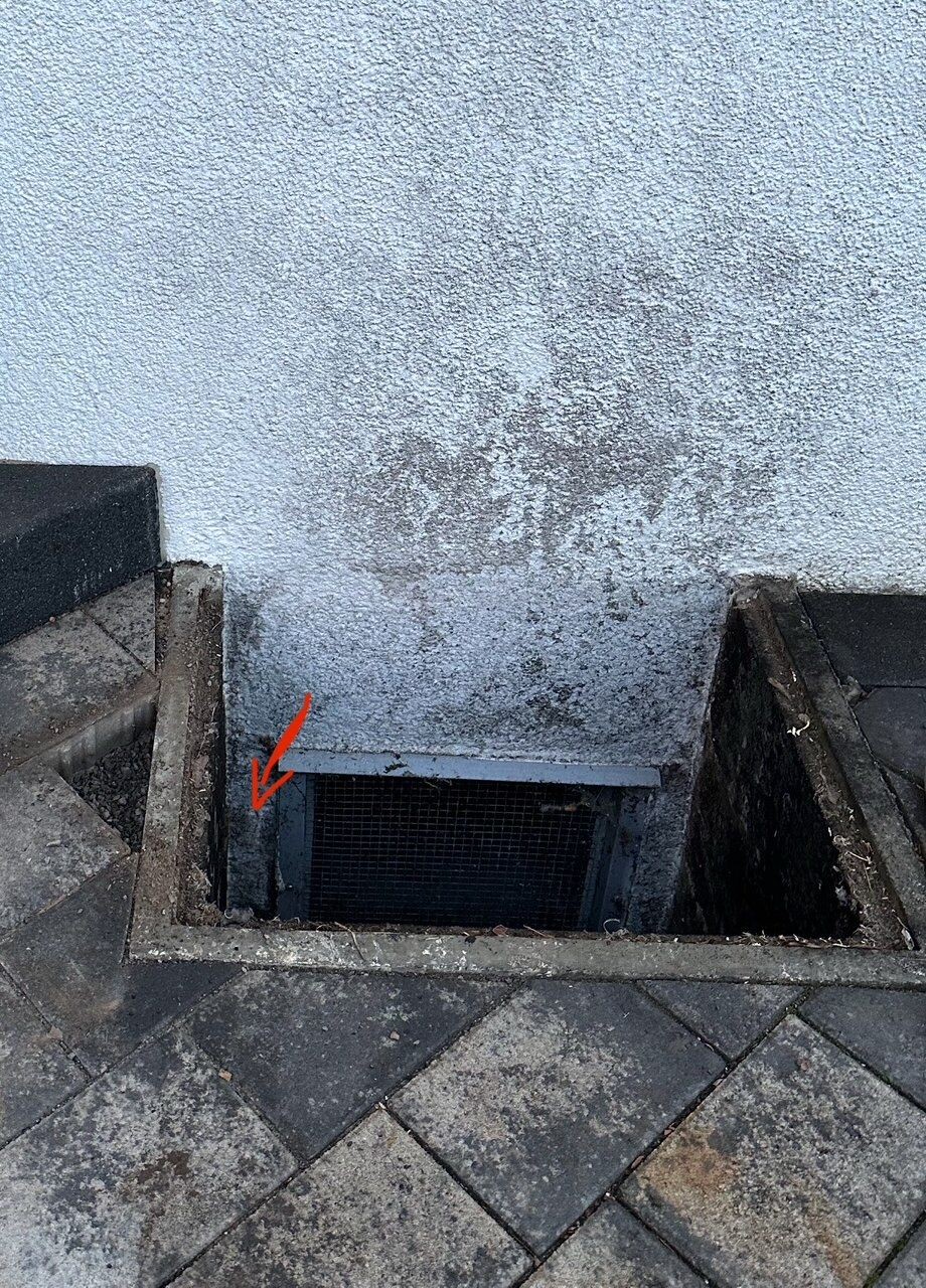 Außenwand mit Lüftungsgitter am Bodenbereich, umgeben von Pflastersteinen.