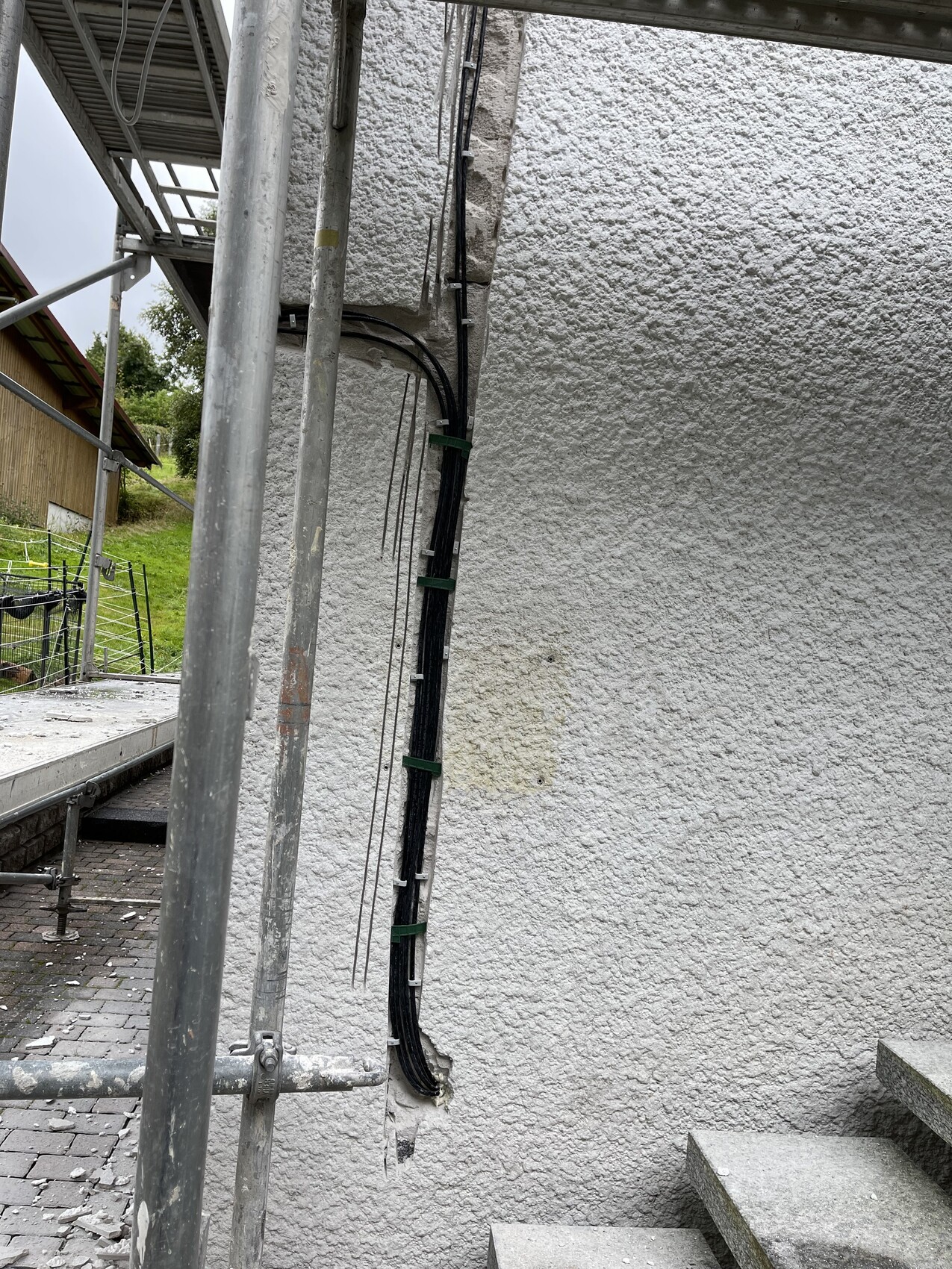 Außenaufnahme: Gerüst links neben einer grob texturierten weißen Wand; Kabelstränge an der Wand.