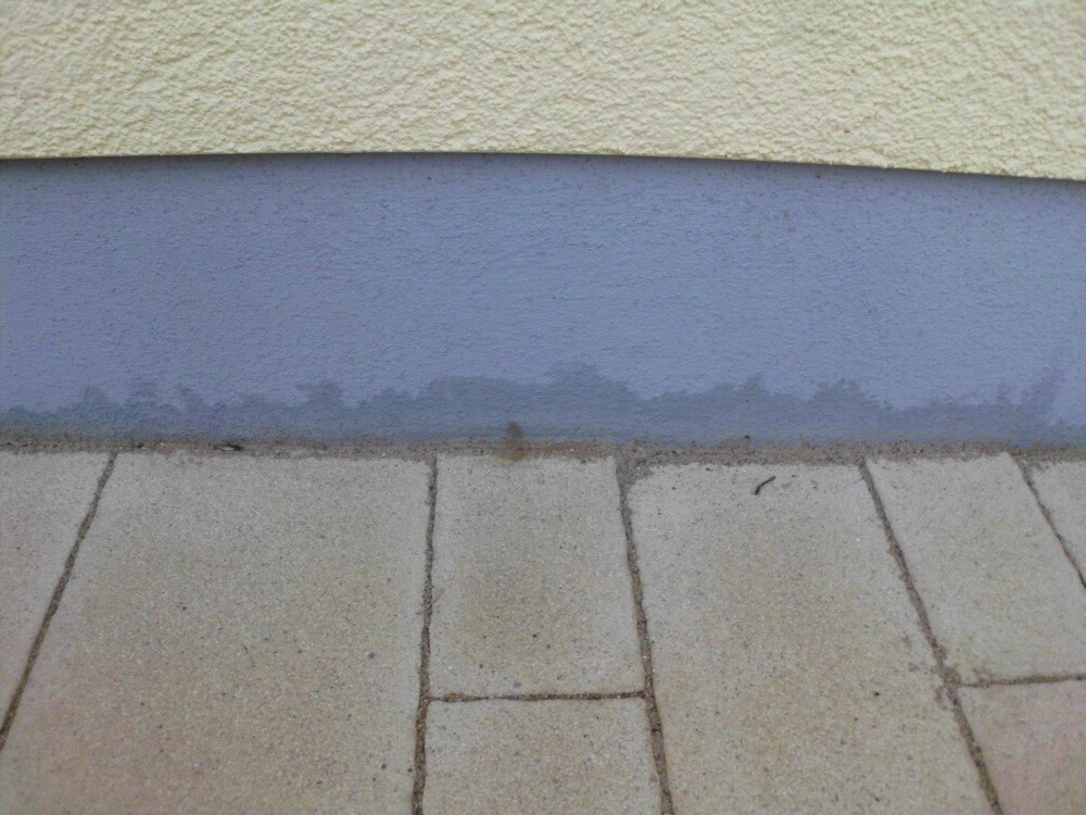Außenwand in gelb mit blauem Streifen am unteren Rand, davor gepflasterter Gehweg