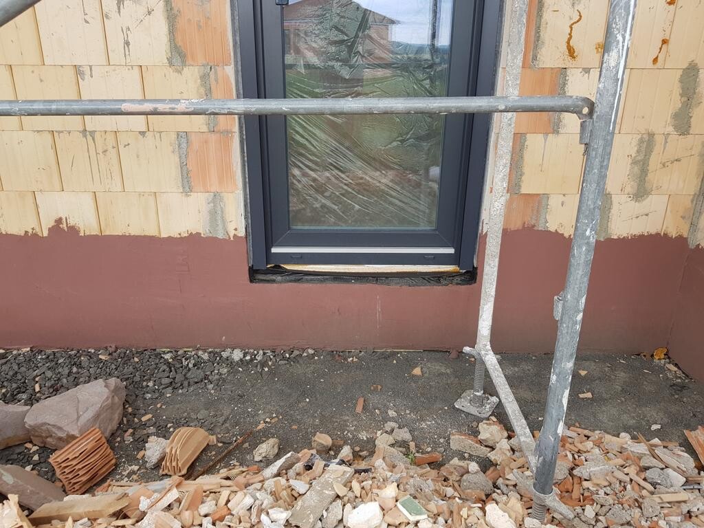 Außenwand unter Bauarbeiten: Fensterrahmen mit Gerüststäben davor, Schutt und Ziegelreste am Boden.