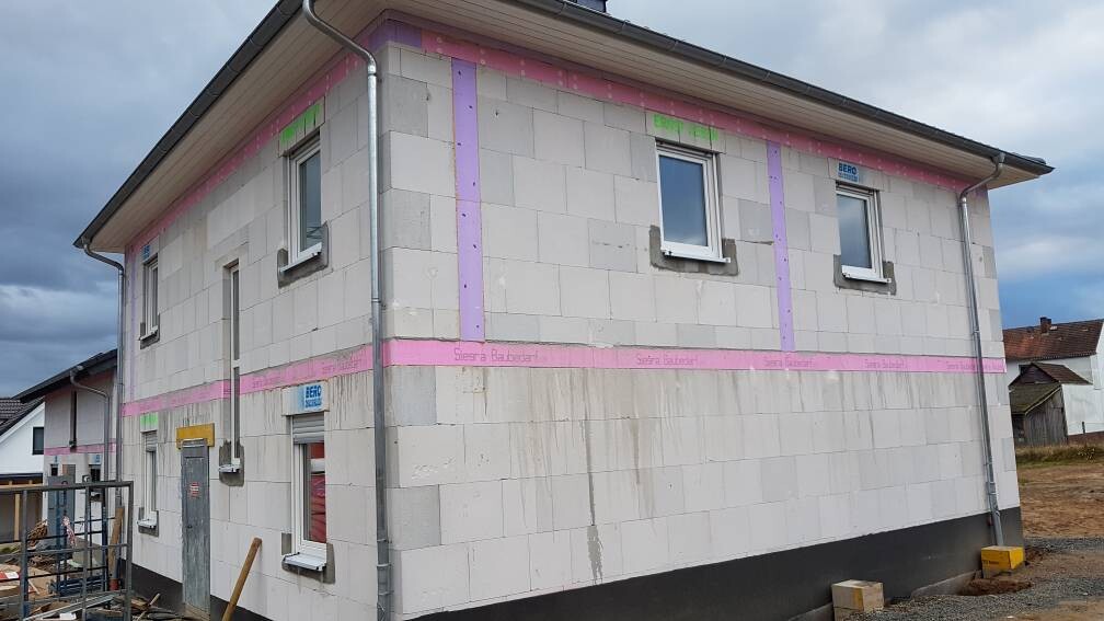 Baustellenhaus mit weißen Dämmplatten, rosa Streifen, zwei Fenster und Baugeräten davor