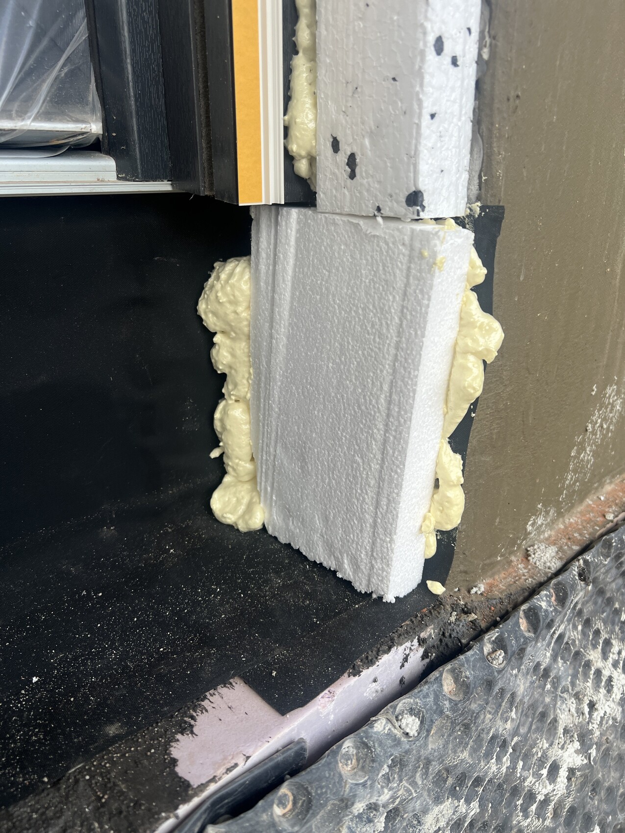Nahaufnahme: Weißer Styroporblock am Fensterrahmen, gelber Expansionsschaum quillt seitlich heraus.