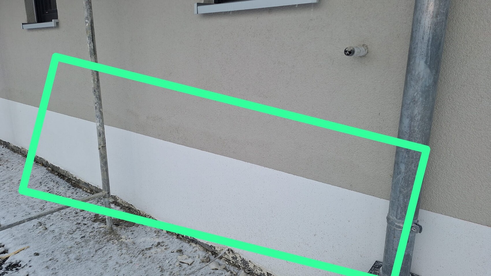 Außenwand mit beigem Verputz, weißem Sockel, Rohre und Gerüstpfosten; grün markiertes Rechteck.