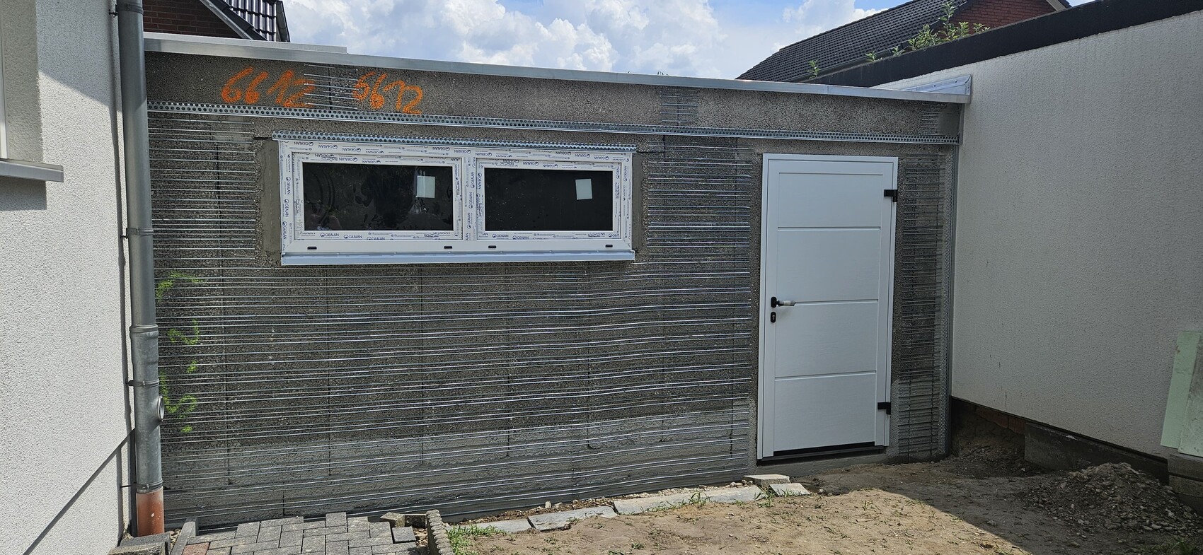 Außenansicht eines Neubaus mit Metallverkleidung, zwei Fenstern und Tür; orange Graffiti.