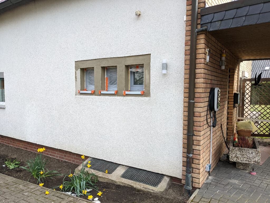 Außenansicht eines Hauses mit weißer Fassade, drei kleine Fenster, Narzissen im Beet.