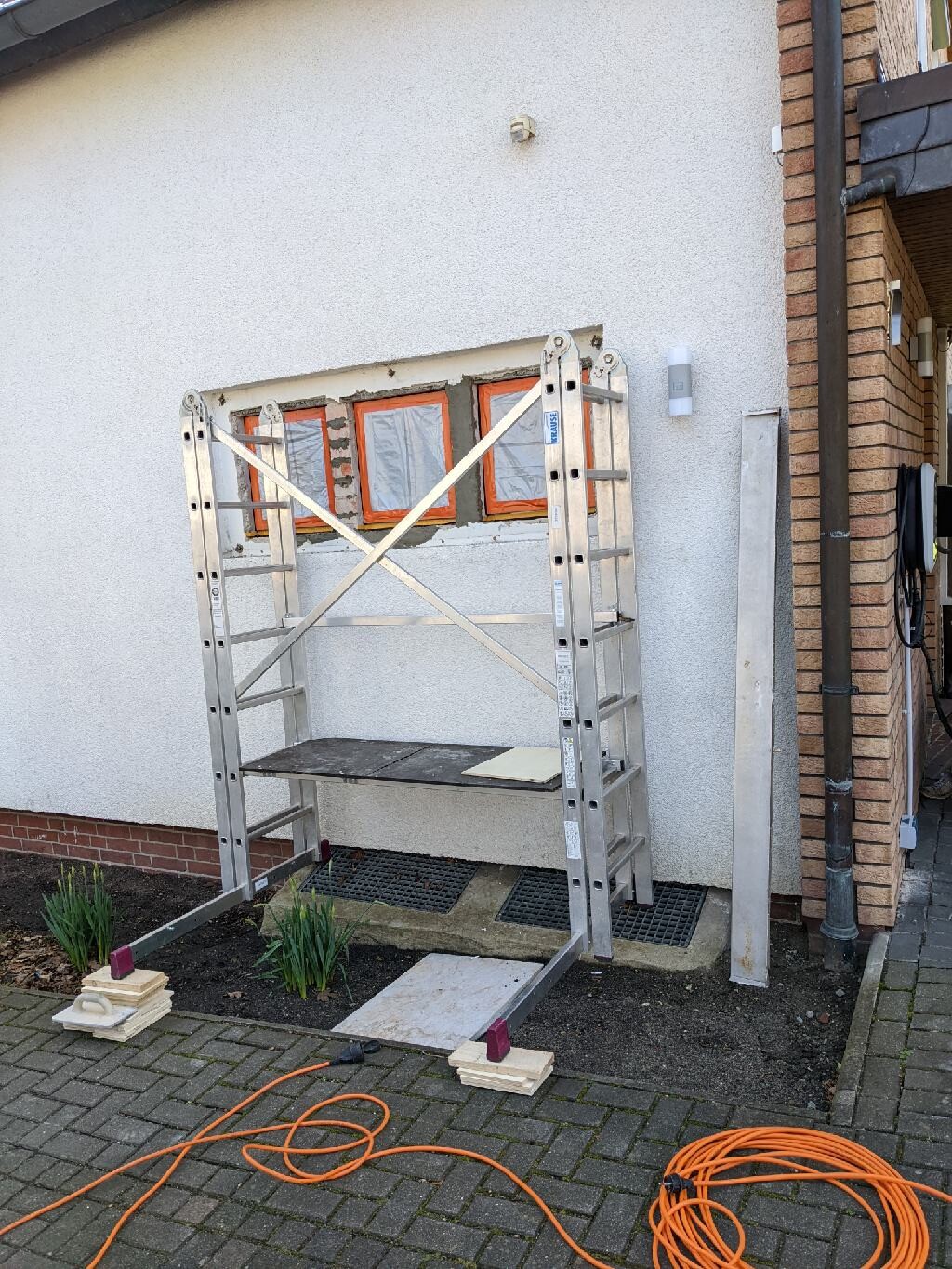 Stahlgerüst vor weißer Hauswand mit orange eingerahmten Fenstern; Kabelrolle liegt auf dem Boden.