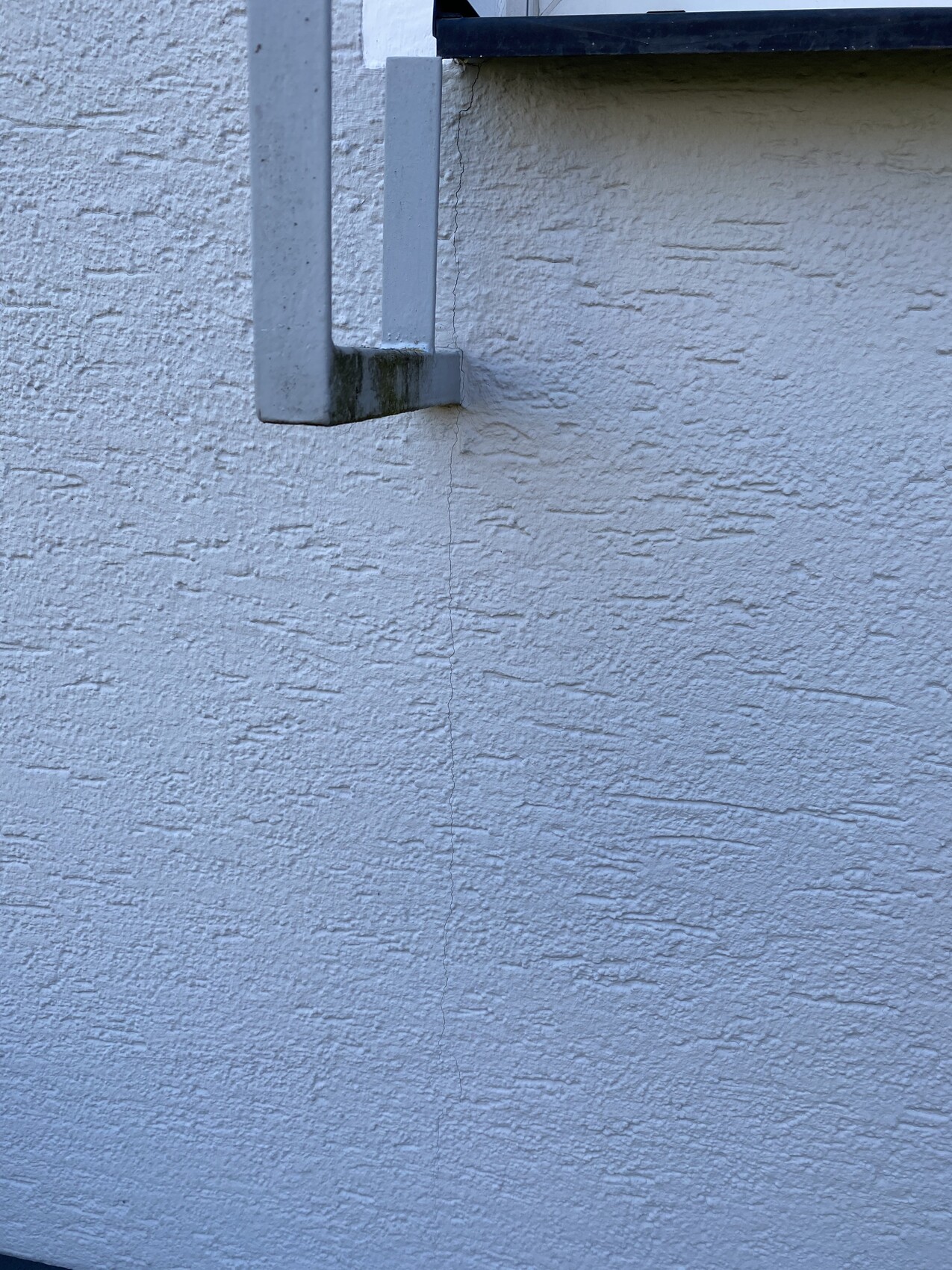 Texturierte weiße Außenwand; graue Metallhalterung an der oberen linken Seite; feiner Wandriss.