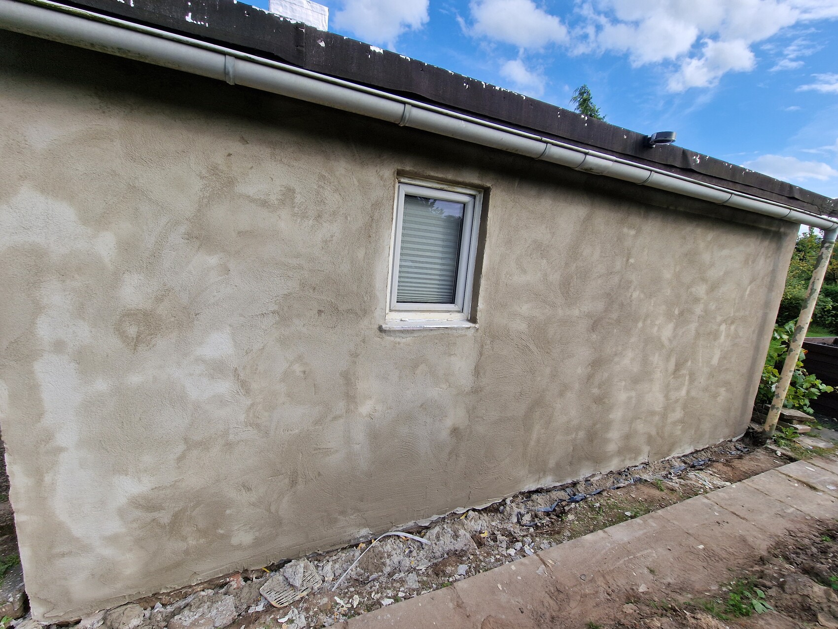 Außenwand eines Gebäudes mit kleinem Fenster, rauem Putz, Dachrinne und Baugrund mit Geröll.