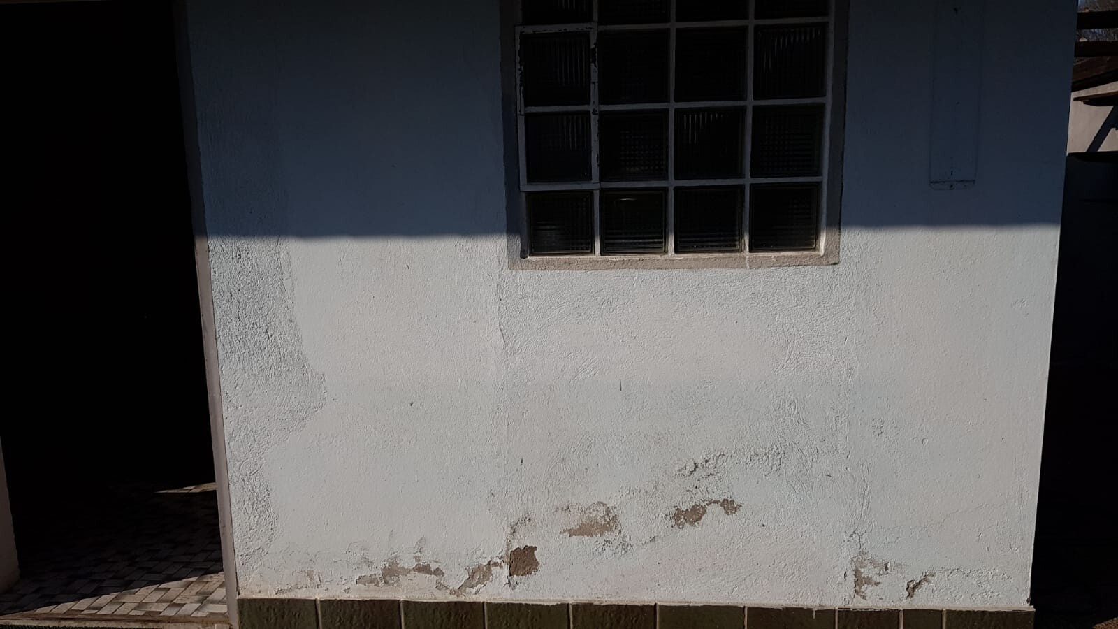 Weiße Außenwand mit kleinem Fenster; Putzrisse und Feuchtigkeitsschäden unten.