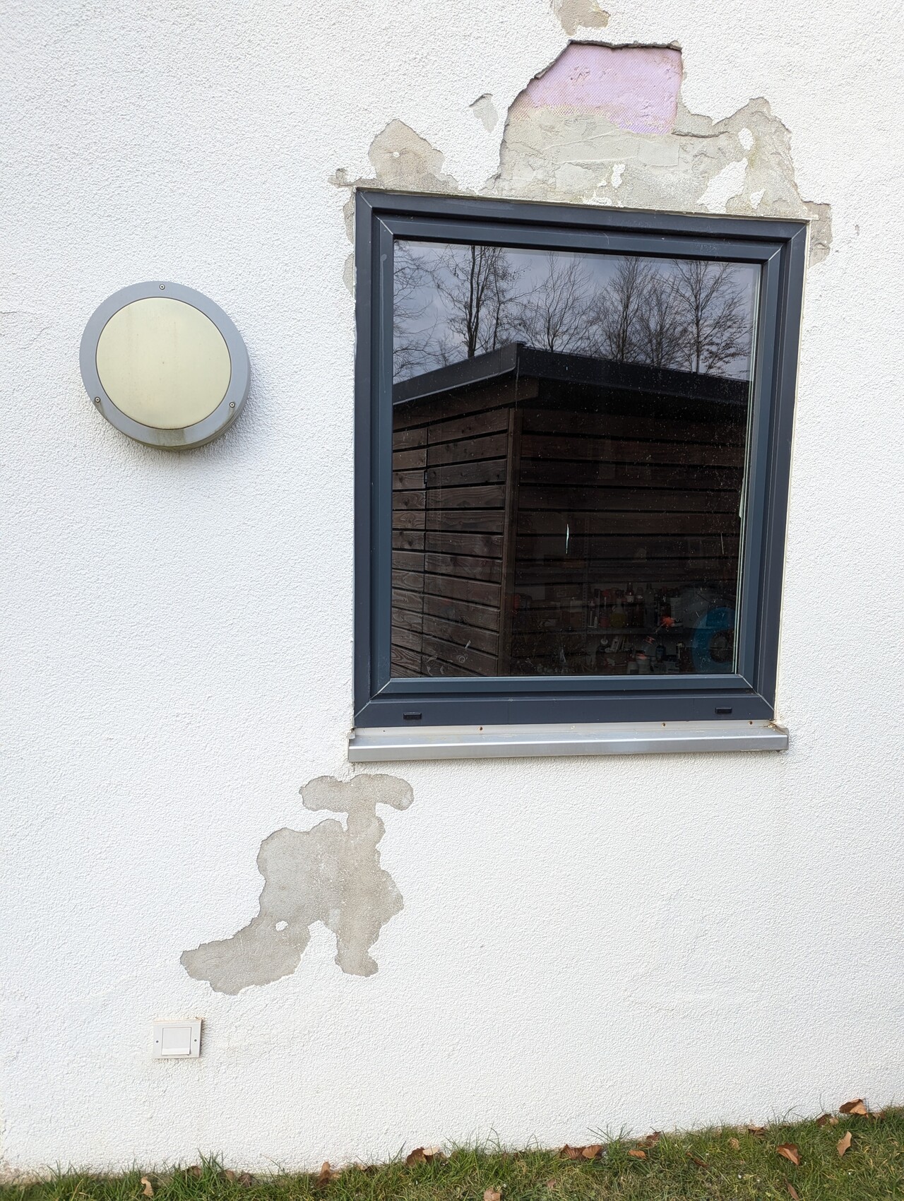 Weiße Außenwand mit Fenster und runder Außenleuchte; Putzabbrüche, Reflektion von Holzhaus.
