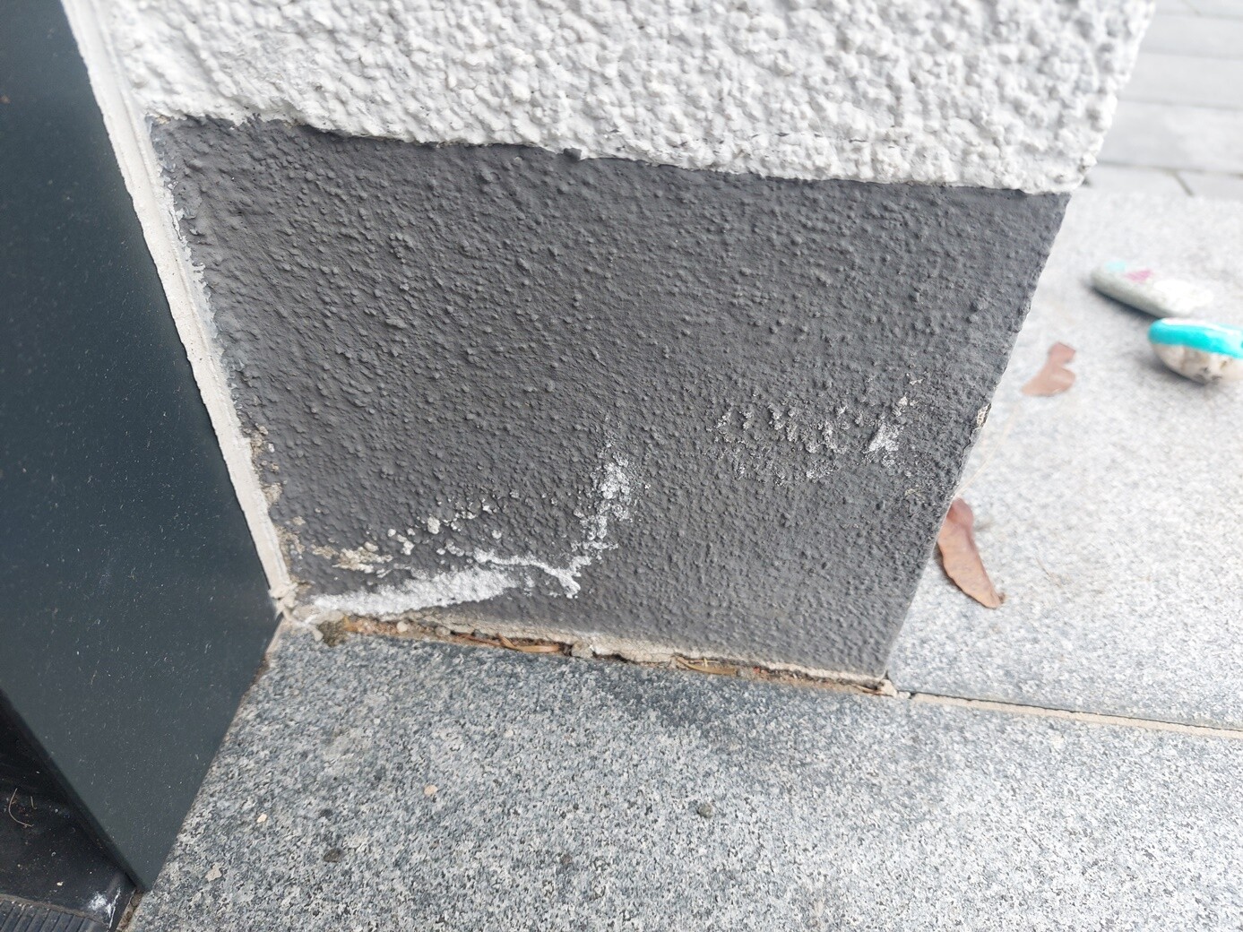 Unterer Teil einer grauen, rauen Putzwand am Gebäudesockel; Pflasterboden mit Blattresten.