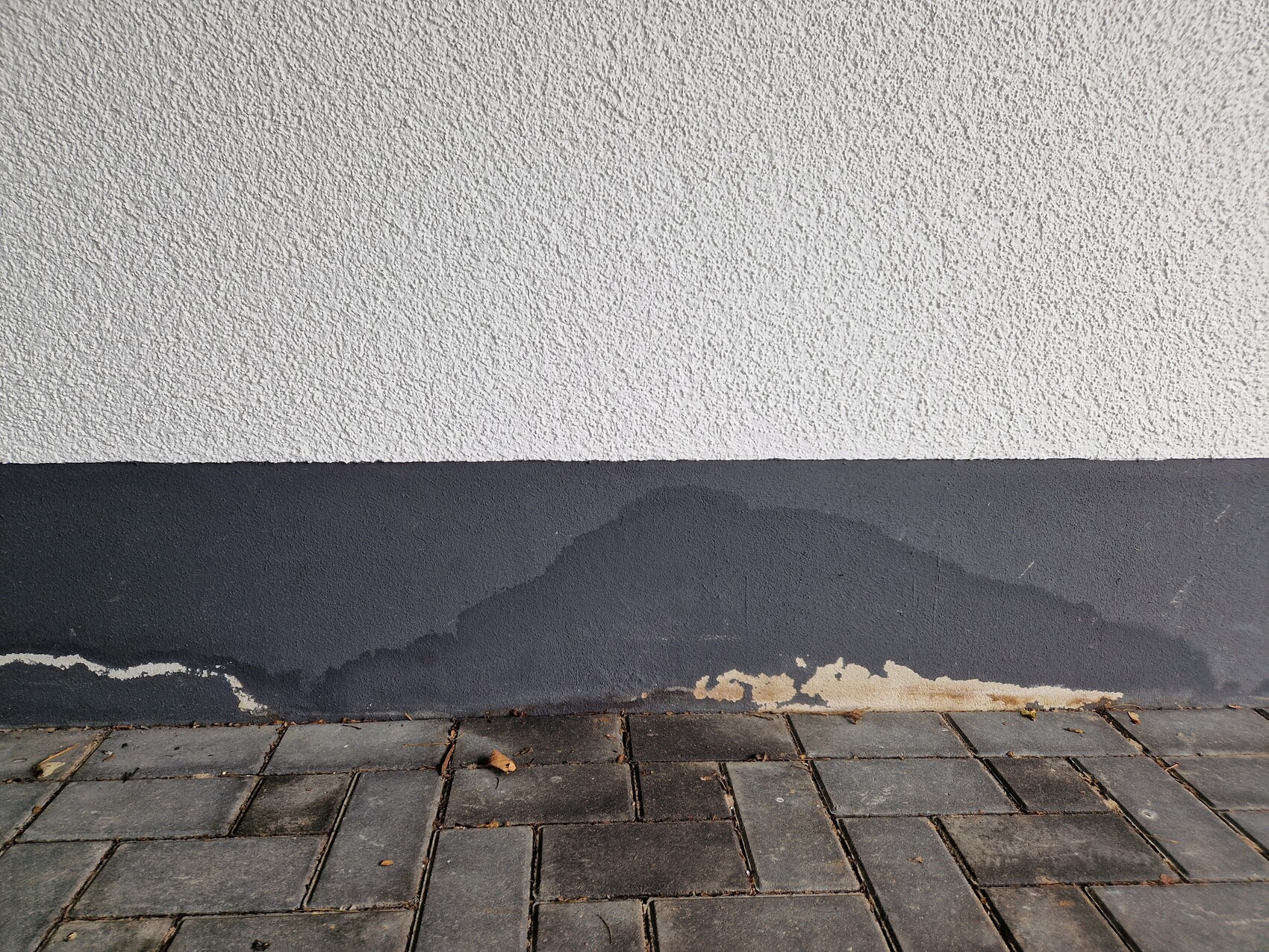 Weiße Putzwand über dunkelgrauem Sockel; Abplatzungen unten, gepflasterter Gehweg davor
