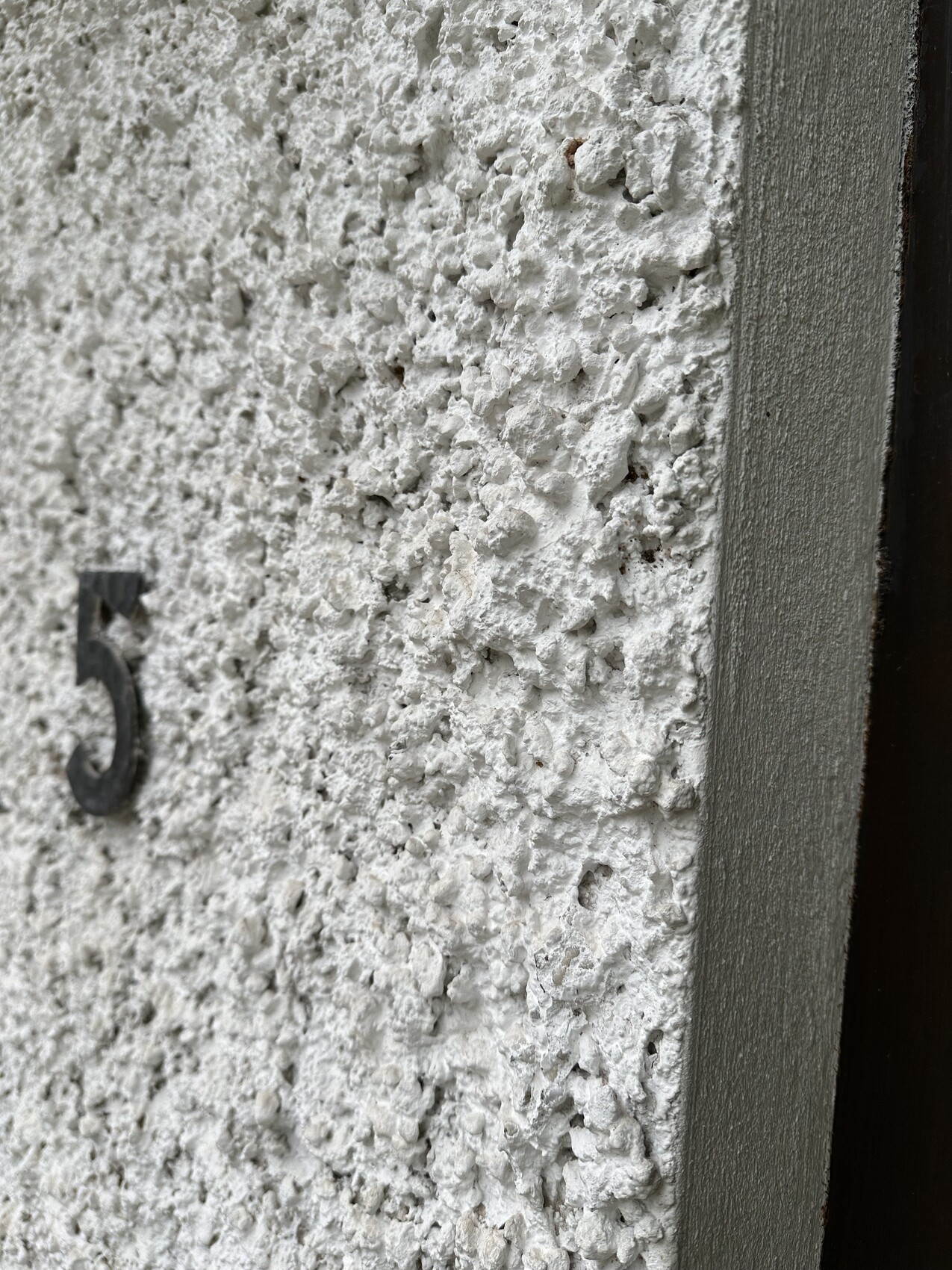 Grobe weiße Rauputz-Wand mit schwarzer Zahl 5; rechter Türrahmen sichtbar.