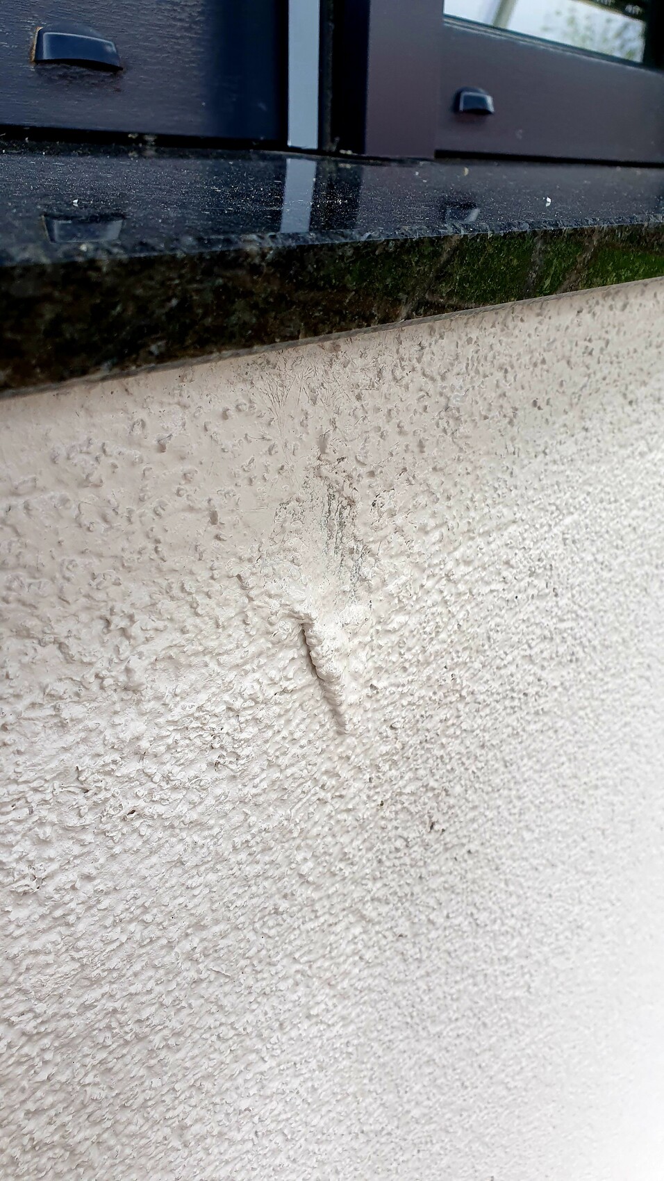 Außenwand mit grober Putzstruktur, vertikaler Putzbruch unter dunklem Fensterbrett.