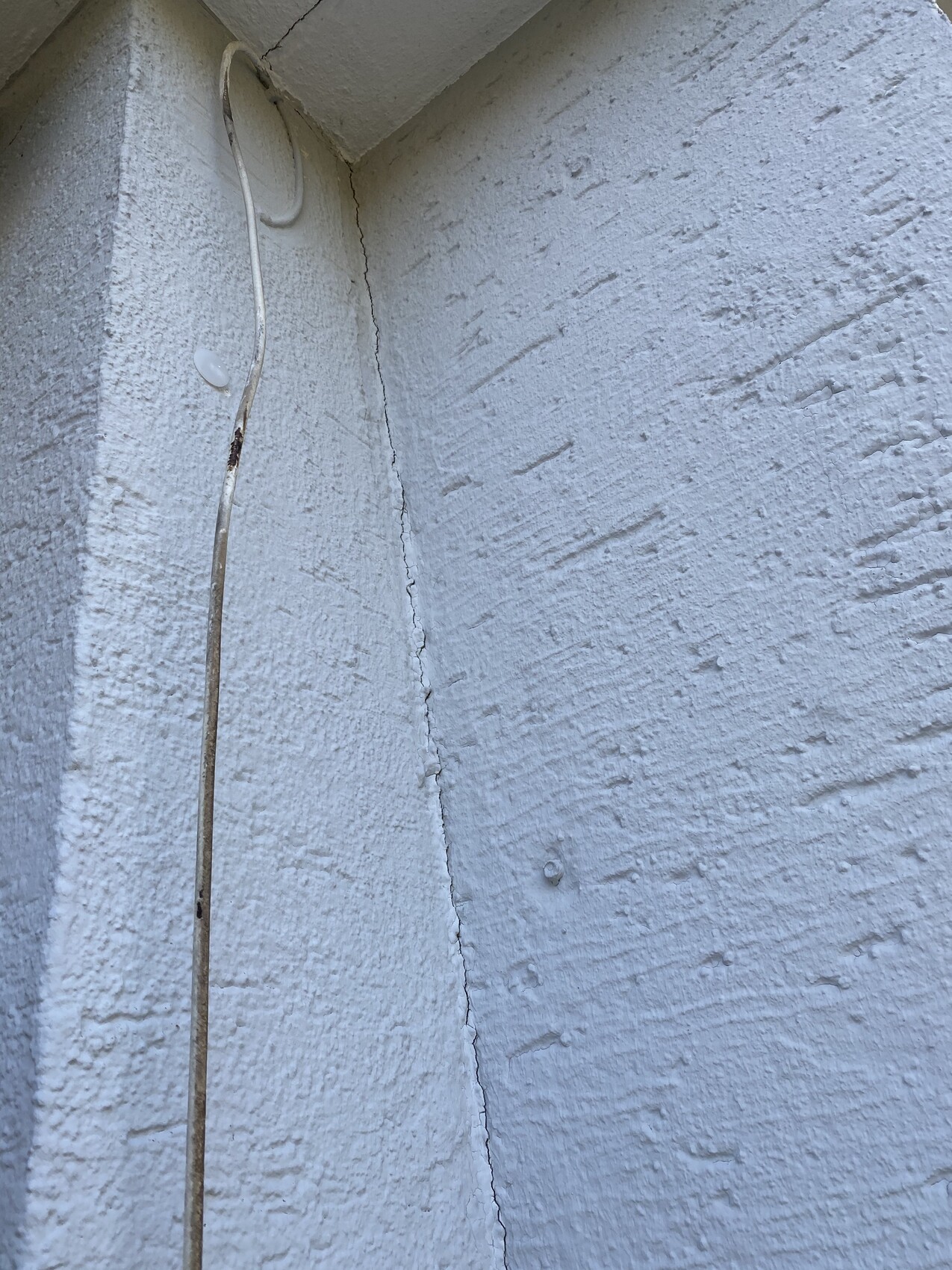 Rauhe weiße Außenwand mit vertikalem Riss; eine lose Leitung hängt an der Wand.