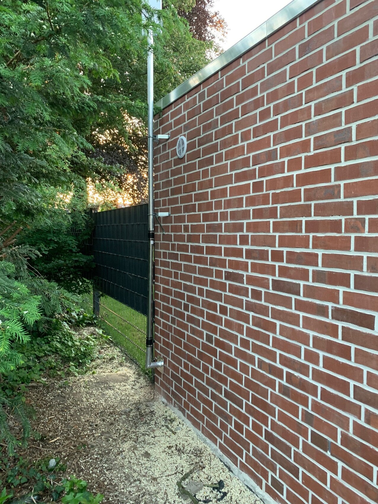 Außenansicht: rote Backsteinwand mit senkrechtem Metallrohr, Lüftungsgitter, Grünpflanzen.