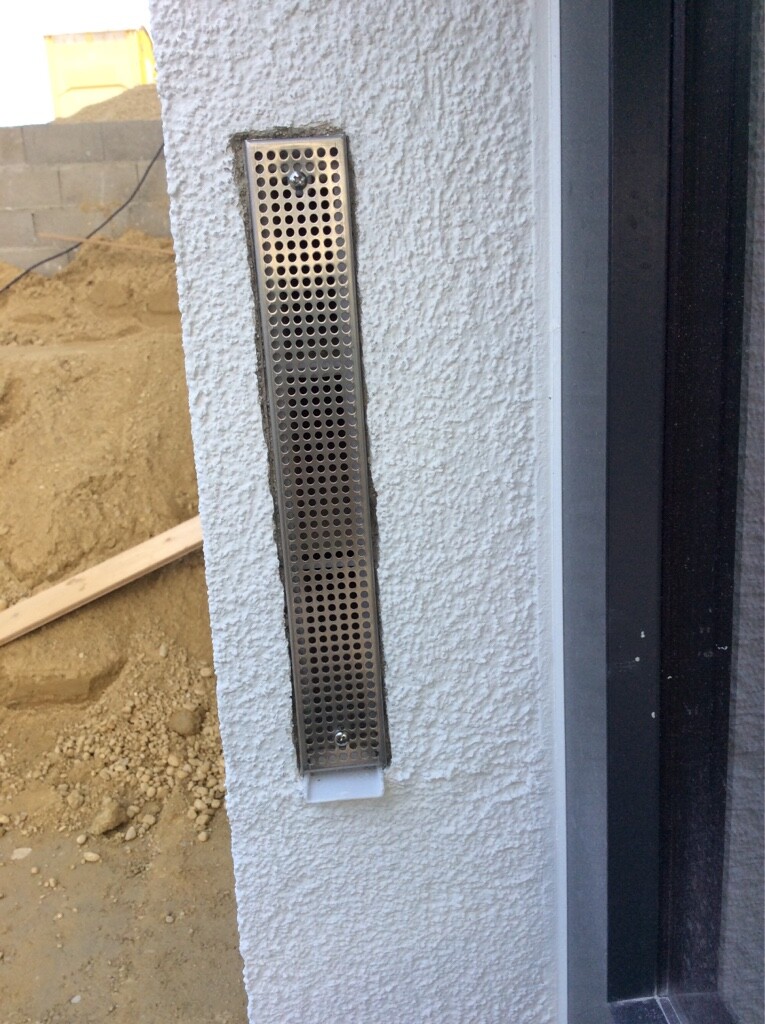 Vertikale Metall-Ventilationsgitter an weißer Außenwand neben Tür; Baustelle im Hintergrund.