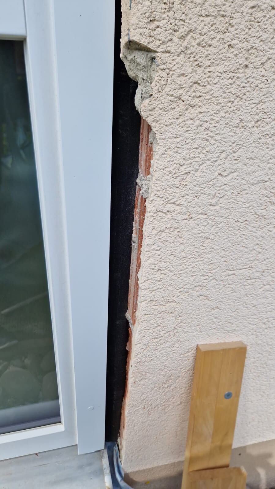 Außenwand neben weißem Fensterrahmen: Putzriss offenbart rotem Backstein, Holzbrett davor.