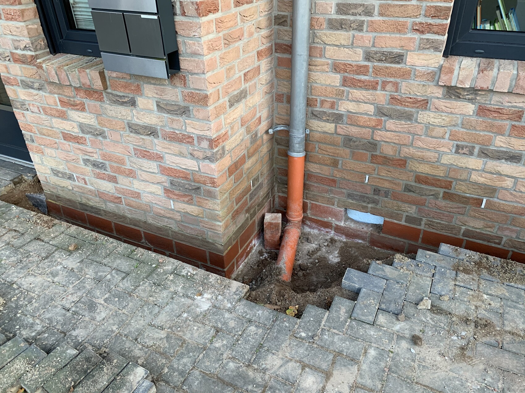 Eckansicht eines Ziegelhauses: graues Abflussrohr trifft auf orangefarbenes Rohr am Fundament.