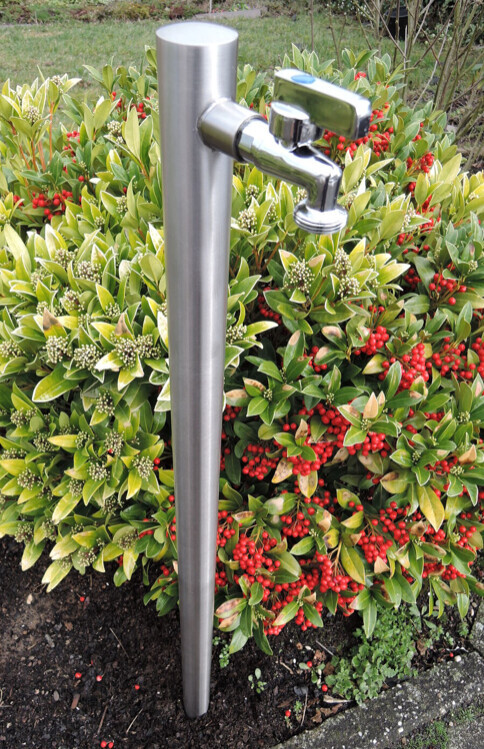 Edelstahl-Wasserhahn an Metallstange, davor Busch mit roten Beeren