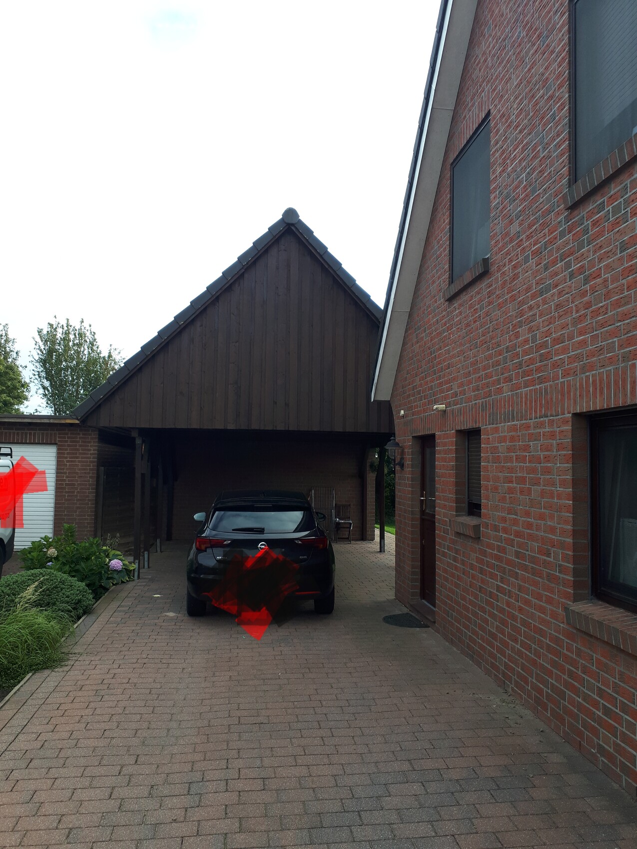 Schwarzes Auto im Carport zwischen braunem Holzvordach und rotem Backsteinhaus, gepflasterte Einfahrt