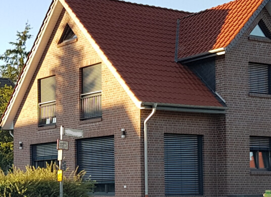 Ziegelhaus mit rotem Satteldach, Fenstern mit Rollläden und Straßenschild.