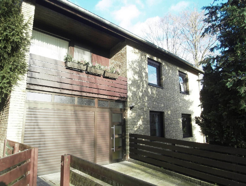 Zweistöckiges Haus aus hellen Ziegeln mit brauner Garagen-Tür, Balkonpflanzen und Holzzäunen.