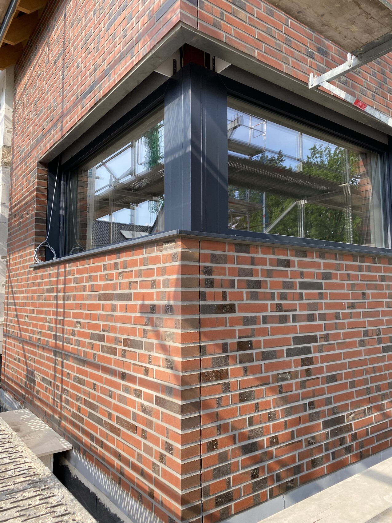 Eckhaus aus roten Ziegeln mit großem Fenster in dunklem Metallrahmen; Baugerüst im Hintergrund.